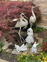 Wide angle photo showing two tall metal bird statues with white feather detail and two smaller statues, a girl and a bunny, in garden space surrounded by plants and a small rock border.