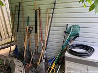 Overview of garden tools including long-handled rakes, shovels, edger, weed puller, pond net, sprayer, cultvator, and coiled hose leaning against house siding.