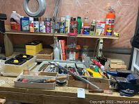 Shows assortment of spray paint, stain, lubricants, hand tools, files, chisels, and hardware arranged on wooden bench surface and shelf above
