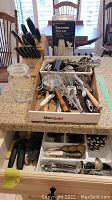 Full view of the lot showing kitchen utensils in a cardboard tray, cutlery drawer with utensils, and a wooden Kitchen Aid knife block on the counter.