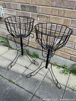 Pair of black metal plant stands against brick wall on paved surface, showing full height and basket shape