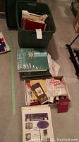 Green plastic storage bin filled with albums, binders and stamp supplies, plus albums and supplement laid out on floor
