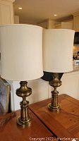 Pair of brass or metal base lamps with tall cylindrical white fabric shades, shown on wooden surface.