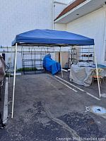 Fully assembled E-Z Up shelter canopy in blue fabric, standing upright in a parking lot with metal frame visible.