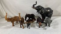 Group photo showing seven elephant figurines of various sizes and materials including wood and a possibly composite black elephant with tusks.