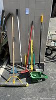 Full set of nine long handled garden tools arranged against a beige shed wall outdoors on asphalt. Includes rakes, brooms, shovels, and a garden scoop with wooden, metal and plastic handles mostly worn and weathered.