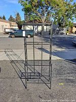 Full front view of black metal plant stand outdoors on pavement with houses, trees, and vehicles in background.