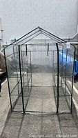 Full front view of assembled portable greenhouse with front zipper door opened halfway, showing the interior wire shelves and metal frame structure.