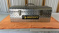Front view of vintage Husky diamond plate toolbox sitting on orange cloth, showing entire toolbox and handle.