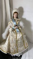 Full view of the doll standing with white embroidered dress, gold trim, blue satin sash, and feather stole.