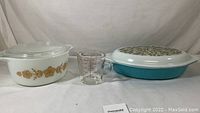 White 1.5 quart Pyrex casserole dish with gold floral design and clear lid, blue 1.5 quart casserole with olive design lid, and clear Pyrex measuring cup with red markings shown together.