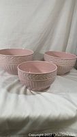 Three pink ceramic mixing bowls of different sizes with Roma decorative pattern, arranged on white fabric background.