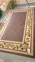 Full view of the rectangular mauve and cream area rug with rose floral border pattern laid flat on floor.