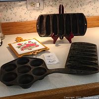 Cast iron ebelskiver pan with seven round molds, two rectangular cornbread molds with eight elongated molds each, and a decorative tile trivet arranged on a countertop.