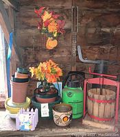 Overall view of various garden and decorative items including the cider press, pots, flowers, and a pump sprayer on a wooden table with a wooden backdrop.
