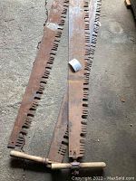 Three long rusty crosscut saw blades laid on garage floor, one with attached wooden handle