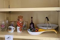 Shelf with assorted European-themed shot glasses, ceramic beer drinking character bottle stopper, glass bell, Mt. Rainier spoonrest, and glass plate with stand.