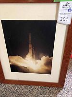 Framed photo capturing a Boeing rocket launch with smoke and flames, wooden frame visible