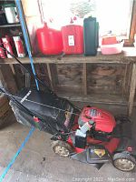 Photo showing the red Troy-Bilt self-propelled lawn mower with black grass collection bag and control handles, placed indoors.