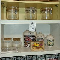 Shelf containing three large glass storage jars with metal clasp lids on top shelf and various jars and decorative items including ceramic jars and wooden box with matchbooks below