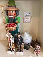 Front view of the 14-inch wooden apple picker nutcracker holding a basket of red apples and a wooden rake, two ceramic Laurel and Hardy figurines, Avon milk glass covered candle shaped like a hen, small ceramic figurines including a woman and a bird, and a pumpkin-shaped ceramic item.