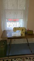 Overall view of Bernina Record sewing machine on laminate table with foldable legs and green carrying case visible on floor.