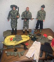 Three male figures standing with various military gear and accessories displayed below on wooden surface.