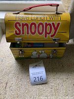 Front view of the Snoopy lunchbox, displaying 'Have Lunch with Snoopy' text and cartoon images on a yellow background with red handle and metal clasps