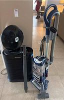 Photo showing the black Honeywell air purifier with a detachable fan unit on top, alongside the blue and gray Shark vacuum and the black Vornado fan.