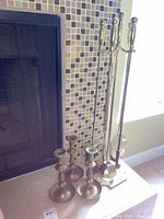 Photo showing brass fireplace tool set with four long handled tools and stand next to fireplace, plus three brass candlesticks.