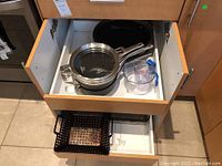 View of Calphalon pots and pans stacked in kitchen drawer with plastic measuring cups and black metal basket below.