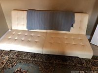 Front view of beige leatherette couch with tufted seat and back cushions, a woven bird and floral throw cover draped over the backrest, sitting on a patterned area rug.