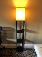 Floor lamp turned on, shows the illuminated beige cloth shade casting warm light and visible bottom shelves.