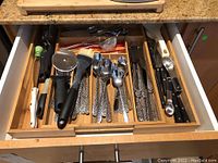 Drawer opened showing organized stainless steel flatware and kitchen utensils including forks, spoons, knives, measuring spoons, spatula, pizza wheel, and strainer.