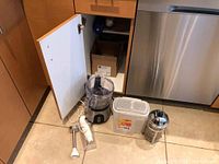 Kitchen items visible inside an open brown cabinet: Hamilton Beach electric food chopper with clear bowl and black base, white electric handheld whisk with cord, translucent plastic container with lid, and stainless steel French press.