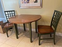 View of the table with two chairs placed on either side, showing the round drop-leaf wood table with green legs and two matching chairs.