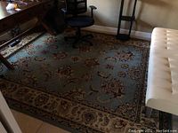 Photo shows the full view of the light blue area rug under office furniture with red and beige floral patterns and bordered design.