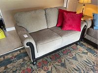 Beige upholstered love seat with 2 red throw pillows on the right side, showing arm, cushions and wood trim on the front and left side