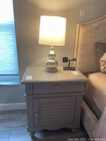 Front view of gray wooden nightstand with drawer and door, base of a bed visible, lamp on top lit.