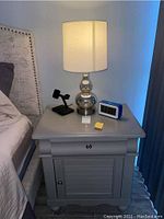 Gray nightstand placed next to a bed with a silver mercury glass lamp, a black stand, and a digital clock on top.