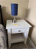White nightstand with one drawer and open shelf on floor beside bed, showing drawer closed and lamp on top.