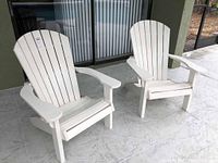 View of two white plastic Adirondack-style patio chairs placed outside on a tiled surface near a building.