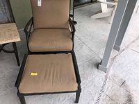 Front-facing photo of one chair with matching ottoman showing brown cushions and black metal frame on patio floor.