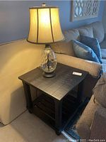 Side table with square black stained wood top and slatted bottom shelf, with clear textured glass lamp on top.