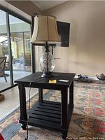 Full view of black solid wood side table with slatted lower shelf and table lamp with clear textured glass base and beige shade sitting on top