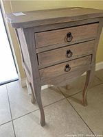 Front view showing three wood drawers with black metal ring pull handles and curved tapered legs.