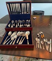 Wide shot showing wooden box with 3 drawer levels containing various forks, knives and spoons, plus additional utensils laid out on the table.