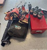 Photo showing three folded red and black camping chairs, a red metal cooler and six green camping gas canisters