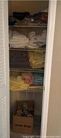 Closet shelves showing neatly folded bath towels in multiple colors, table linens folded and stacked, and a box of toilet paper rolls underneath the shelves.