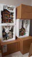 Stacked ceramic Christmas village buildings in original white foam packaging and cardboard boxes, showing detailed painted holiday buildings with wiring for lighting.